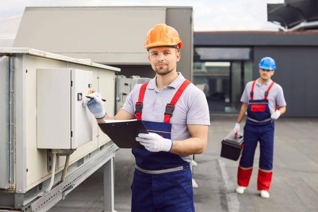 Two men in overalls stand beside an air conditioner, preparing for thermal inspections in a mold removal service.